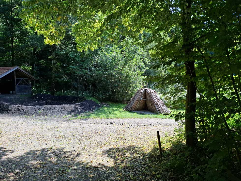 Waldlichtung mit Köhlerstelle, rechts steht aufgeschichtetes Holz als Pyramide, links der Meiler